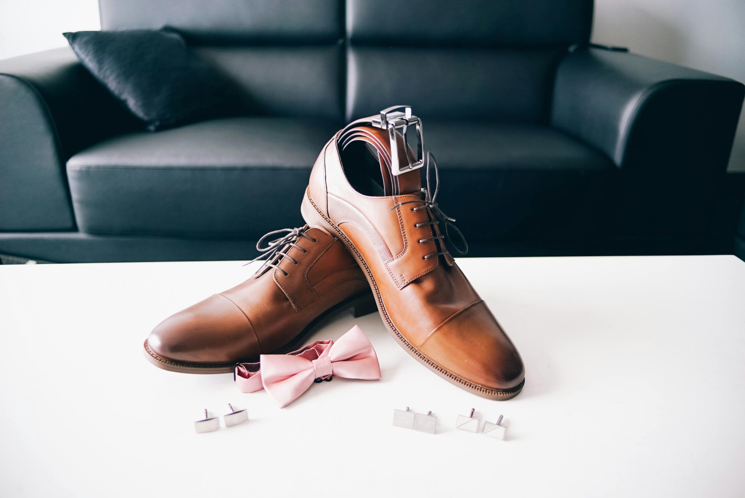 Stylish men's brown dress shoes with a pink bow tie and cufflinks placed on a white table, perfect for formal events.